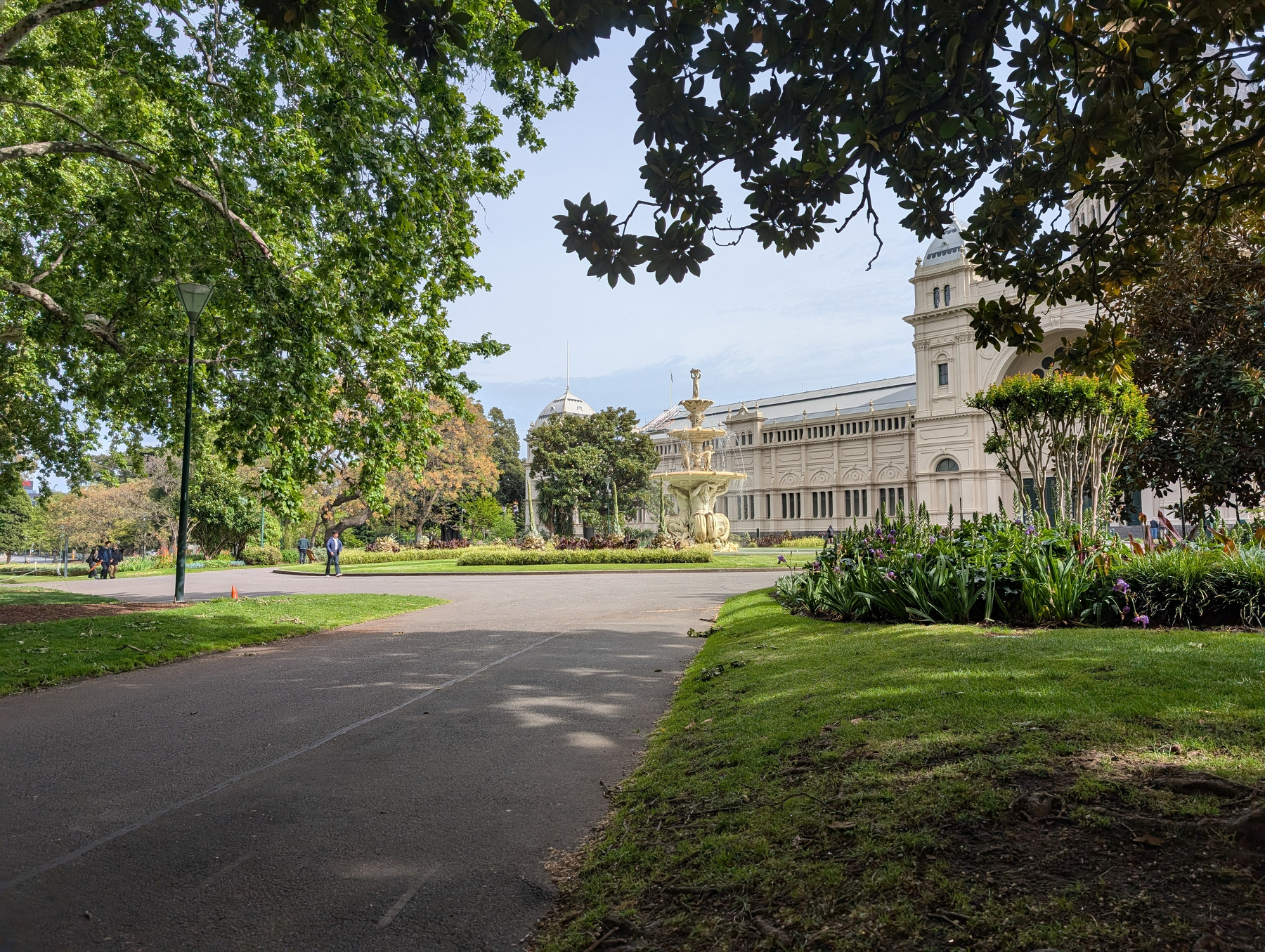 Auto-generated description: A wide pathway bordered by lush greenery leads to an ornate fountain and a historical building under a partly cloudy sky.