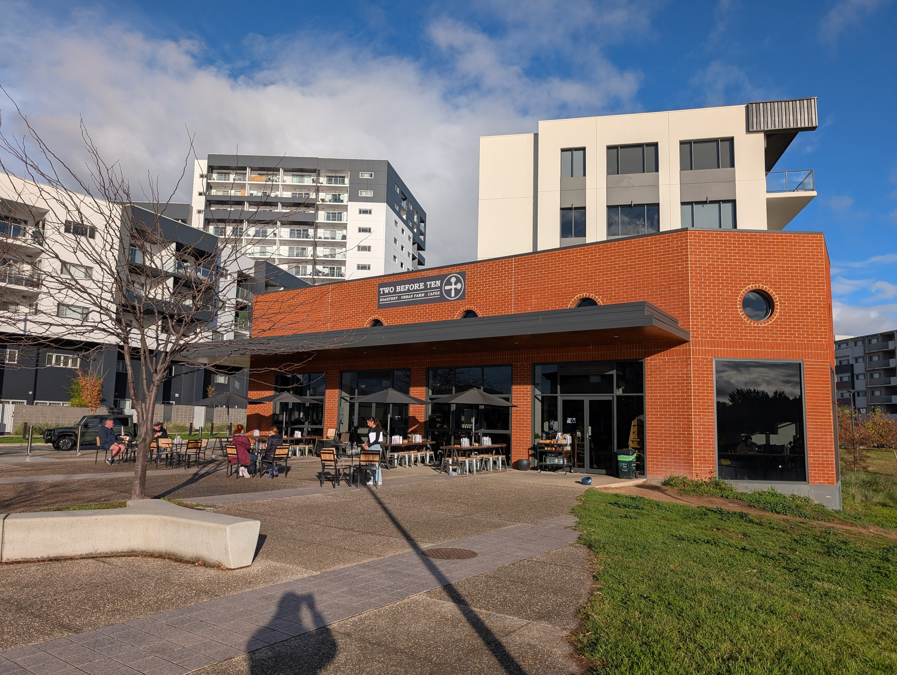Auto-generated description: A brick café with outdoor seating is located in front of modern residential buildings under a partly cloudy sky.