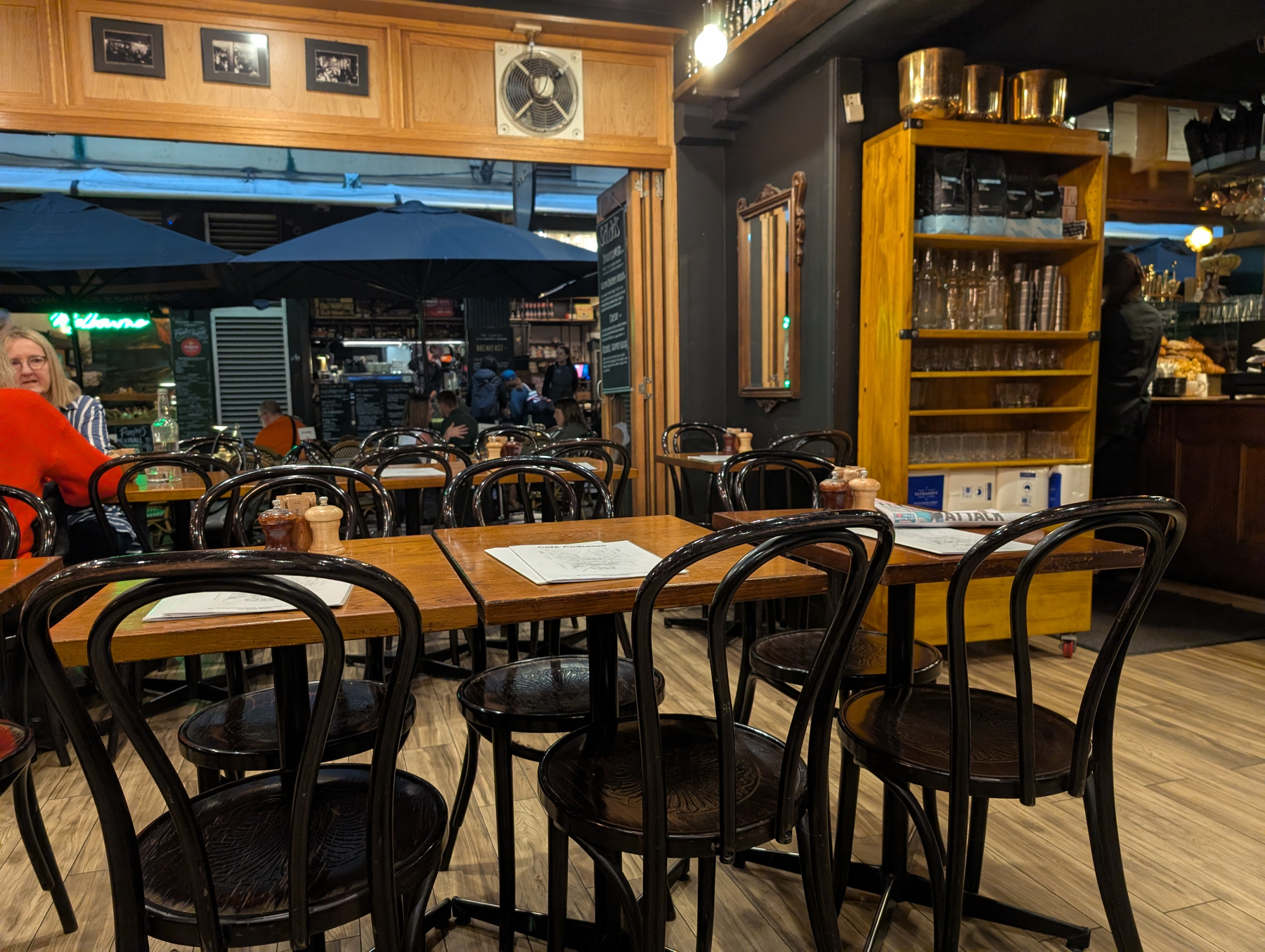 A cozy cafe setting with wooden tables and chairs, a shelving unit filled with glassware, and outdoor seating visible through an open doorway.