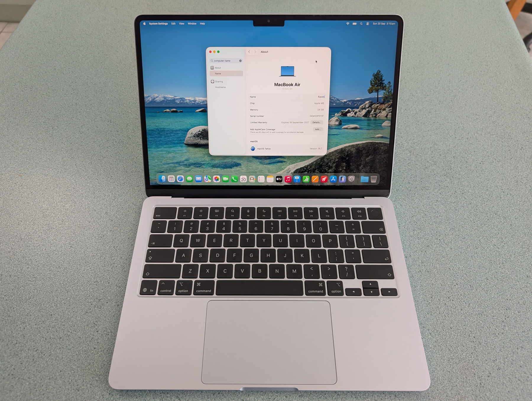 Auto-generated description: A MacBook Air laptop is open on a light-colored surface, displaying system information on the screen with a scenic beach wallpaper in the background.